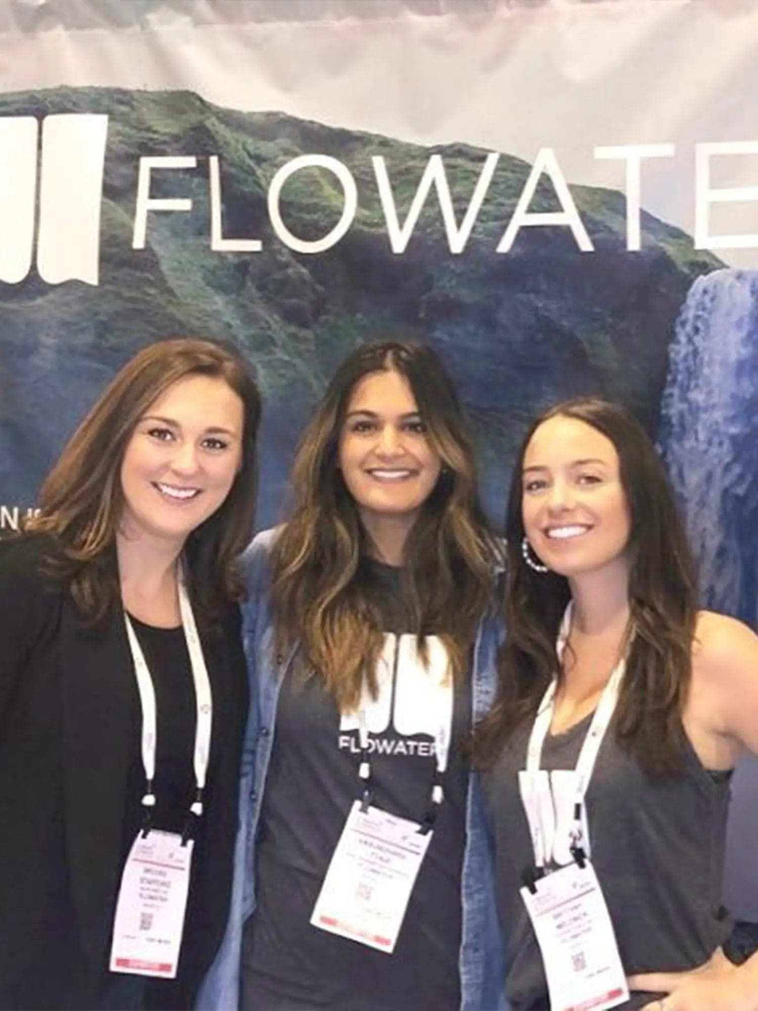 Three women wearing conference badges stand in front of a Flowater banner, smiling at the camera.