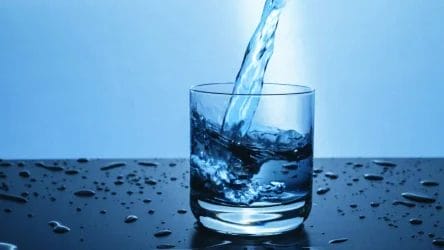 A clear glass is being filled with water, with water splashing inside and droplets scattered on the dark surface around the glass.