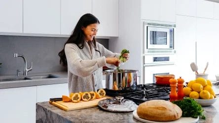 A woman adds greens to a pot on a stove in a modern kitchen, with bread, cut squash, lemons, and other ingredients on the counter.