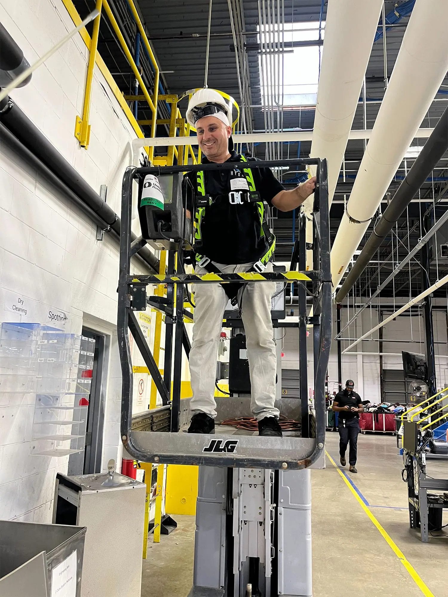 A worker wearing safety gear stands on a raised JLG lift inside an industrial facility with pipes and equipment visible in the background.