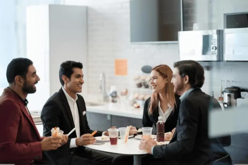 Four people in business attire sit around a small table in an office kitchen, eating lunch and talking, with beverages and food containers on the table.